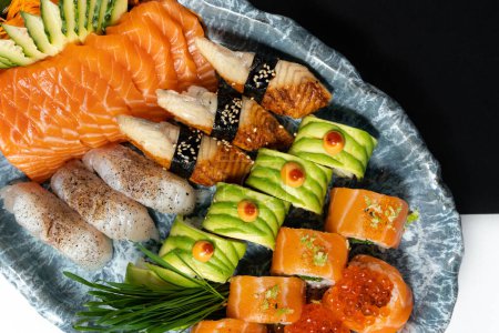 Téléchargez les photos : Assortiment de sushis en rouleaux avec avocat, anguille, saumon, caviar rouge, pétoncle dans une assiette ovale en céramique avec un motif bleu sur fond de papier noir et blanc. À côté se trouve une figurine en plâtre. - en image libre de droit