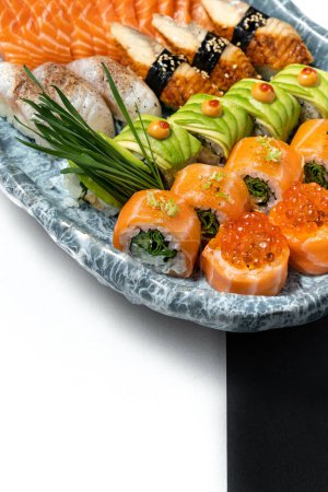 Photo for Assorted sushi set of rolls with avocado, eel, salmon, red caviar, scallop in an oval ceramic plate with a blue pattern on a black and white paper background. Next to it is a plaster figurine. - Royalty Free Image