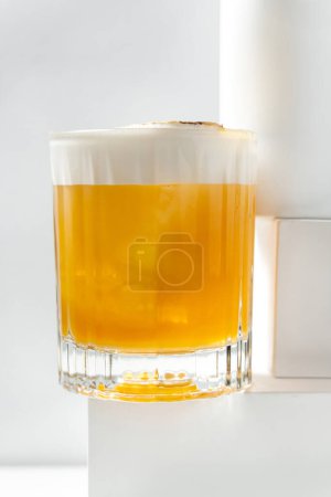 Photo for Mango Sour cocktail in a transparent glass with a piece of ice and citrus foam, topped with banana and mango powder. The cocktail stands on a plaster figure among other figures on a white background. - Royalty Free Image