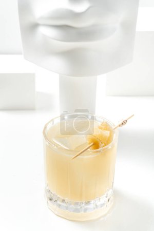Téléchargez les photos : Citrus Cocktail aigre dans un verre transparent avec des morceaux de miel sur une brochette. Le cocktail se tient sur un fond blanc parmi les figures de plâtre. - en image libre de droit