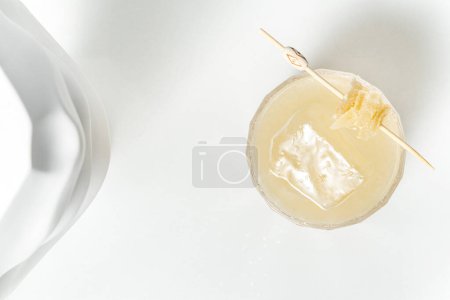 Téléchargez les photos : Citrus Cocktail aigre dans un verre transparent avec des morceaux de miel sur une brochette. Le cocktail se tient sur un fond blanc parmi les figures de plâtre. - en image libre de droit