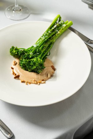 Téléchargez les photos : Choux de brocoli grillés avec sauce aux noix et cacahuètes dans une assiette en céramique légère. Le plat se tient sur une nappe légère, à côté d'un verre de vin blanc et de couverts. - en image libre de droit