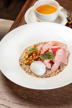 Photo for Quinoa with ham and poached egg and vegetable tabouleh in a light ceramic plate. The dish stands on a brown tabletop, next to it is a cup and a transparent teapot with tea. - Royalty Free Image