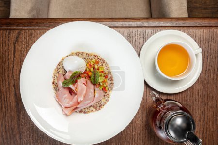 Photo for Quinoa with ham and poached egg and vegetable tabouleh in a light ceramic plate. The dish stands on a brown tabletop, next to it is a cup and a transparent teapot with tea. - Royalty Free Image
