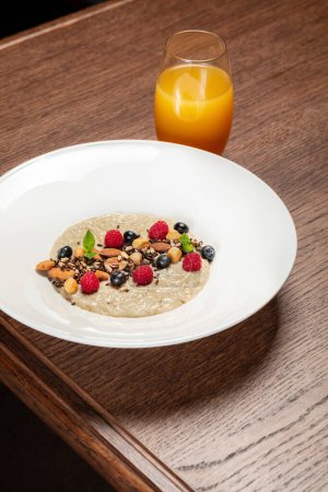 Photo for Oatmeal with nuts of various kinds, raspberries and blueberries with mint leaves in a light ceramic plate. The dish stands on a wooden table, next to a glass with orange juice. - Royalty Free Image