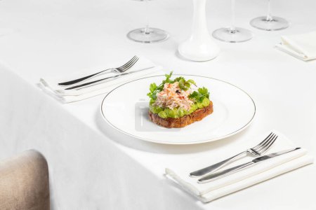 Photo for Bruschetta with poached egg, avocado puree, lettuce and parsley with crabmeat on top. Food on a light ceramic plate, on a table with a light tablecloth. Nearby is a vase and cutlery. - Royalty Free Image