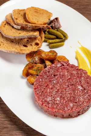 Photo for Veal tartare with rye bread toasts, fried potatoes, pickled cucumbers and anchovies with mustard sauce. Food lies on a light plate on a brown wooden tabletop. - Royalty Free Image