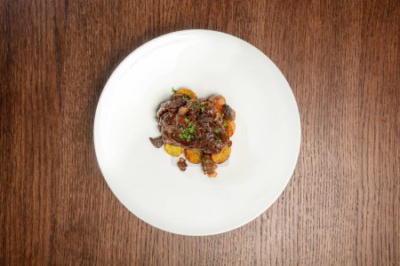 Photo for Veal cheeks in wine sauce with chopped herbs on top and fried potatoes with cribs on a light ceramic plate. The plate stands on a brown, wooden countertop. - Royalty Free Image