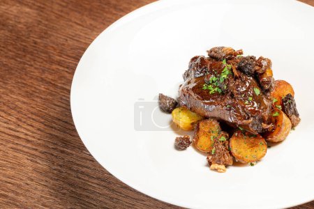 Photo for Veal cheeks in wine sauce with chopped herbs on top and fried potatoes with cribs on a light ceramic plate. The plate stands on a brown, wooden countertop. - Royalty Free Image