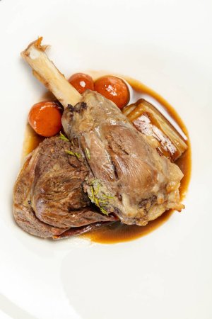 Photo for Leg of lamb baked with wine and tomatoes and wine sauce in a light ceramic plate. The plate stands on a wooden, brown countertop. - Royalty Free Image
