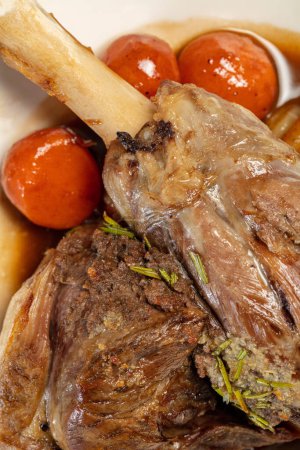 Photo for Leg of lamb baked with wine and tomatoes and wine sauce in a light ceramic plate. The plate stands on a wooden, brown countertop. - Royalty Free Image
