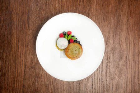 Photo for White chocolate fondant with pistachio cream inside, vanilla ice cream, pistachio chips, blueberries and raspberries, mint leaves nearby. Dessert on a light plate on a wooden tabletop. - Royalty Free Image