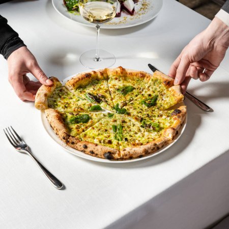 Photo for Pizza with sourdough dough, four cheeses with jalapenos and pistachios and parmesan cheese on top on a light ceramic plate. The dish stands on a light tablecloth and two hands take a piece from different sides. - Royalty Free Image