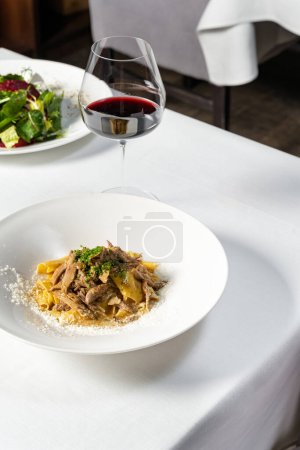 Photo for Homemade pasta with rabbit in a creamy sauce, chopped green onions and Parmesan cheese on top. The food lies in a light ceramic plate on a white tablecloth, next to it is a glass of red wine. - Royalty Free Image