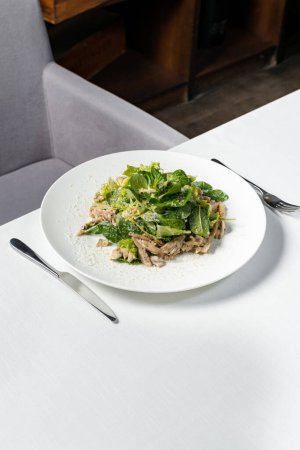 Téléchargez les photos : Mélanger la salade avec la poitrine de poulet et le parmesan dans une assiette en céramique légère. L'assiette est sur une nappe légère, à côté de laquelle se trouve un verre de vin blanc et des couverts. - en image libre de droit