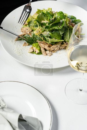 Photo for Mix salad with chicken breast and Parmesan cheese on a light ceramic plate. The plate is on a light tablecloth, next to it is a glass of white wine and cutlery. - Royalty Free Image