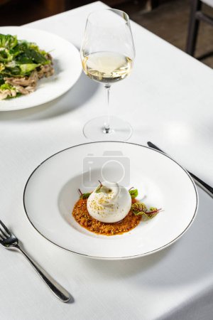 Photo for Buffalo mozzarella with lettuce. vegetable stew, nuts and wine sauce. The food lies in a light ceramic plate. The dish sits on a light tablecloth, next to a glass of white wine and cutlery. - Royalty Free Image