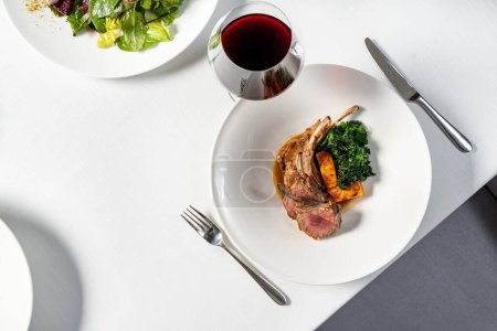 Photo for Rack of lamb grilled and cut into portions, next to it lies a piece of grilled pumpkin and fried lettuce. The food lies in a light ceramic plate on a light tablecloth, next to a glass of red wine. - Royalty Free Image