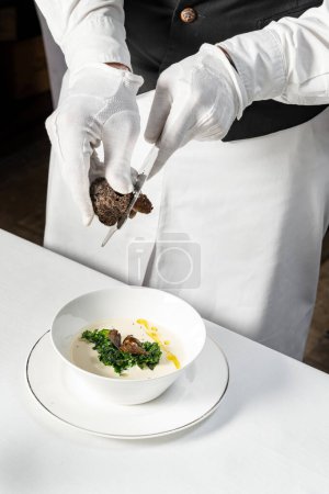 Photo for Mushroom cream soup with truffle slices, olive oil and herbs, in a light ceramic soup bowl on a saucer. The dish stands on a light tablecloth, the waiter rubs the truffle into a plate. - Royalty Free Image
