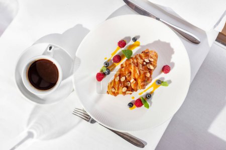 Photo for French croissant with caramel and hazelnut on top, next to it on a plate are raspberries and blueberries with sauce and mint leaves. Food on a light plate on a table with a light tablecloth, a cup of coffee nearby. - Royalty Free Image