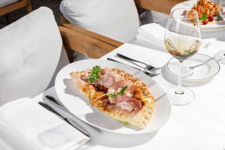 Photo for Calzone pizza with mozzarella cheese, slices of ham and basil leaves on top. The dish lies on a light plate. Taoelka stands on the tablecloth, next to it is a glass of white wine and cutlery. - Royalty Free Image