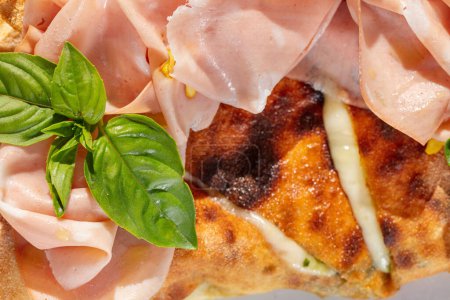 Téléchargez les photos : Pizza Calzone avec fromage mozzarella, tranches de jambon et feuilles de basilic sur le dessus. Le plat repose sur une plaque lumineuse. Taoelka se tient sur la nappe, à côté est un verre de vin blanc et couverts. - en image libre de droit