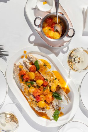Photo for Trout fillet with red and yellow cherry tomatoes in sauce with white wine and basil leaves and Italian herbs in a light plate, next to a glass of white wine and a piala with tomatoes in sauce. - Royalty Free Image