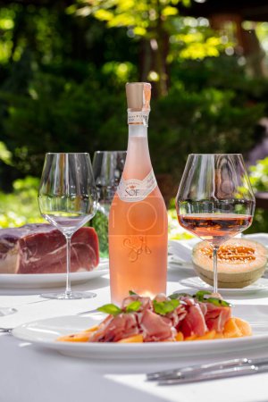 Photo for Salad with melon and prosciutto, basil leaves and balsamic dressing. Nearby is a bottle of rose wine, a glass of rose wine. In the background is a plate with half a melon, a plate with a piece of jamon meat. Dishes are on a table with a white tablecl - Royalty Free Image