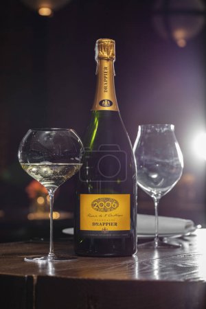Téléchargez les photos : Une bouteille de champagne Grande Sendre 2006 Magnum et une coupe avec du champagne versé à côté. La bouteille et le support en verre sur la table en bois. - en image libre de droit