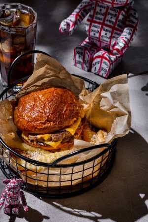 Téléchargez les photos : Hamburger à double côtelette de viande, mayonnaise et fromage, dans un panier en fer avec des frites sur une table en béton avec un verre de cacao cola et jouets en plastique - en image libre de droit