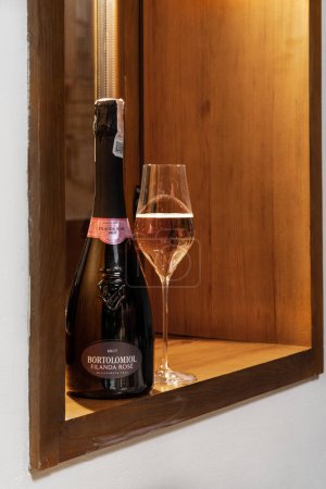 Photo for Bottle and a glass of Bortolomiol Filanda Rose Brut ros sparkling wine stand on a wooden table against a light stone wall with Ukrainian ornaments and wooden window frames. - Royalty Free Image