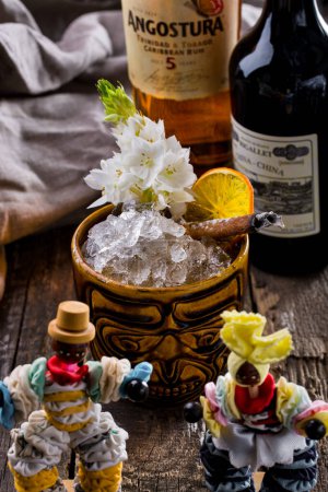 Photo for Rum in a glass with ice with flowers, lemon and cinnamon standing on a wooden table with bottles of rum and a cloth in the background - Royalty Free Image