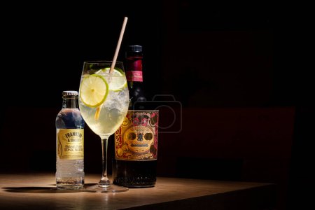 Téléchargez les photos : Un cocktail avec vermouth, tonique, citron, glace et une paille dans un verre qui est sur la table avec une bouteille de tonique et une bouteille de vermouth - en image libre de droit