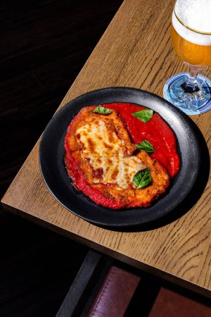 Foto de Chuleta de pollo con queso, albahaca y salsa en un plato sobre la mesa con un vaso de cerveza - Imagen libre de derechos