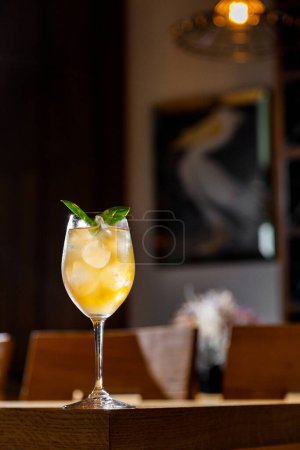 Photo for A cocktail with ice, syrup and basil in a glass that stands on the edge of a wooden table surrounded by a restaurant - Royalty Free Image
