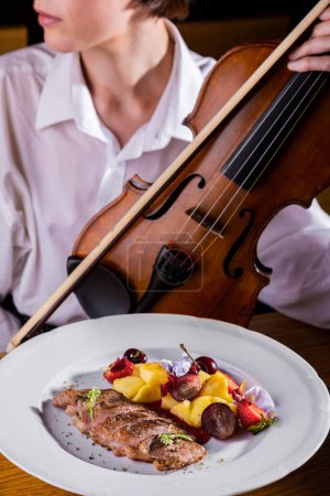 Téléchargez les photos : Morceaux de viande frite avec pommes de terre, raisins, pétales de fleurs, fraises et assaisonnements dans une assiette sur la table sur le fond d'une femme tenant un violon - en image libre de droit