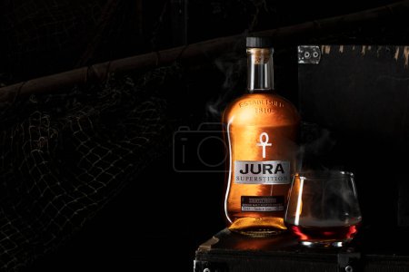 Photo for Jura superstition Whiskey bottle with glass and smoke inside standing on wooden crates with mesh on background - Royalty Free Image