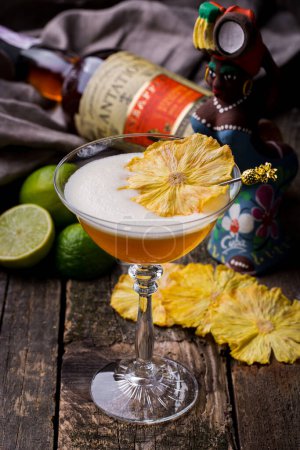 Photo for A cocktail with dried pineapple in a glass standing on a wooden table with a lime, a bottle of wine and a cloth in the background - Royalty Free Image