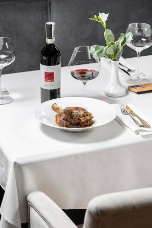 Photo for Leg of lamb baked with wine and tomatoes and wine sauce in a light ceramic plate. The plate stands on a light tablecloth, next to it is a glass and a bottle of red wine, a vase and cutlery. - Royalty Free Image