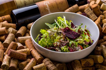Photo for Salad with cheese, arugula, beetroot, endive, tomatoes, sorrel, lettuce and basil in a plate lying on a set of wine corks with wine bottles - Royalty Free Image