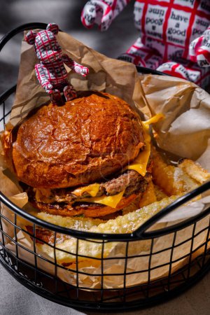 Foto de Hamburguesa con chuleta doble de carne, mayonesa y queso, en una canasta de hierro con papas fritas sobre una mesa de hormigón con un vaso de cola de cacao y juguetes de plástico - Imagen libre de derechos