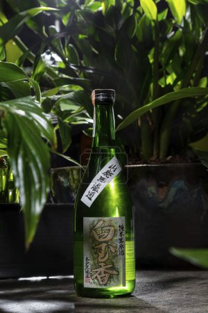 Photo for A bottle of Japanese rice vodka - sake. Sake stands on a gray stone background, tropical plants and a backdrop of gray stone background are arranged around. Nearby stands a wine, transparent glass on a high leg with white wine. - Royalty Free Image