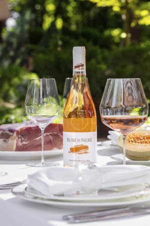 Foto de On the served table, there is a bottle of rose wine, next to it is a glass with poured rose wine. Nearby are plates and glasses. Dishes and wine are on a table with a white tablecloth next to a flower bed with plants. - Imagen libre de derechos