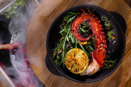 Photo for Fried octopus with thyme, rosemary, lemon, pea sprouts and flower petals in a frying pan that stands on a wooden stand with smoke and a photo frame on the background - Royalty Free Image
