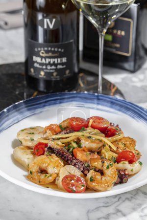Photo for Pasta with seafood. Homemade pasta with scallop, octopus, shrimp and cherry tomatoes in a creamy herb sauce. Pasta lies in a round, light ceramic plate. The plate stands on a light marble table. Nearby is a glass of prosecco, cutlery on a linen napki - Royalty Free Image