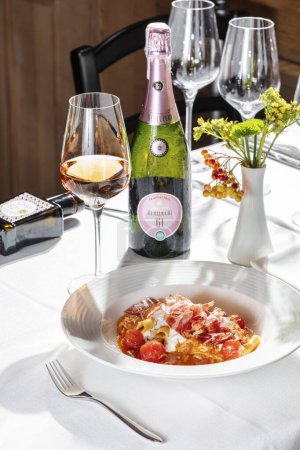 Photo for Pappardelle pasta with prosciutto and stewed tomatoes in wine and cream sauce. Food in a light ceramic plate on a table with a light tablecloth. Nearby is glass of ros, a bottle of Berlucchi '61 Franciacorta Ros, a vase and cutlery. - Royalty Free Image