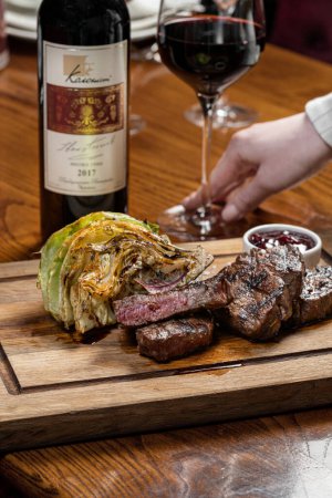 Photo for Three pieces of filet mignon steak grilled, lie on a wooden board. Nearby lies grilled cabbage with balsamic sauce and there is a bowl with cherry sauce. The blackboard lies on a brown table. - Royalty Free Image