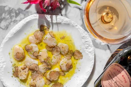 Photo for Scallop carpaccio with truffle mushroom slices in olive oil and herbs. Nearby is a glass of ros sparkling wine and a bottle of sparkling wine. Food in a light ceramic plate on marble countertop. - Royalty Free Image