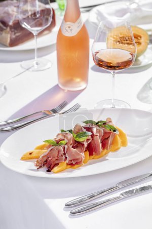 Téléchargez les photos : Salad with melon and prosciutto, basil leaves and balsamic dressing. Nearby is a bottle of rose wine, a glass of rose wine. In the background is a plate with half a melon, a plate with a piece of jamon meat. Dishes are on a table with a white tablecl - en image libre de droit