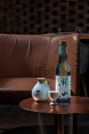 Téléchargez les photos : Une bouteille de saké japonais avec des verres et une carafe est assise sur une table en cuivre à côté d'une chaise en cuir marron. - en image libre de droit